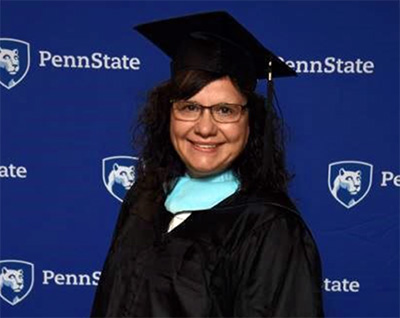 A woman wearing a black graduation cap and gown stands in front of a blue and white background. 