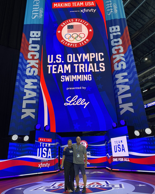 Two people pose in front of electronic screens with red, white, and blue images.