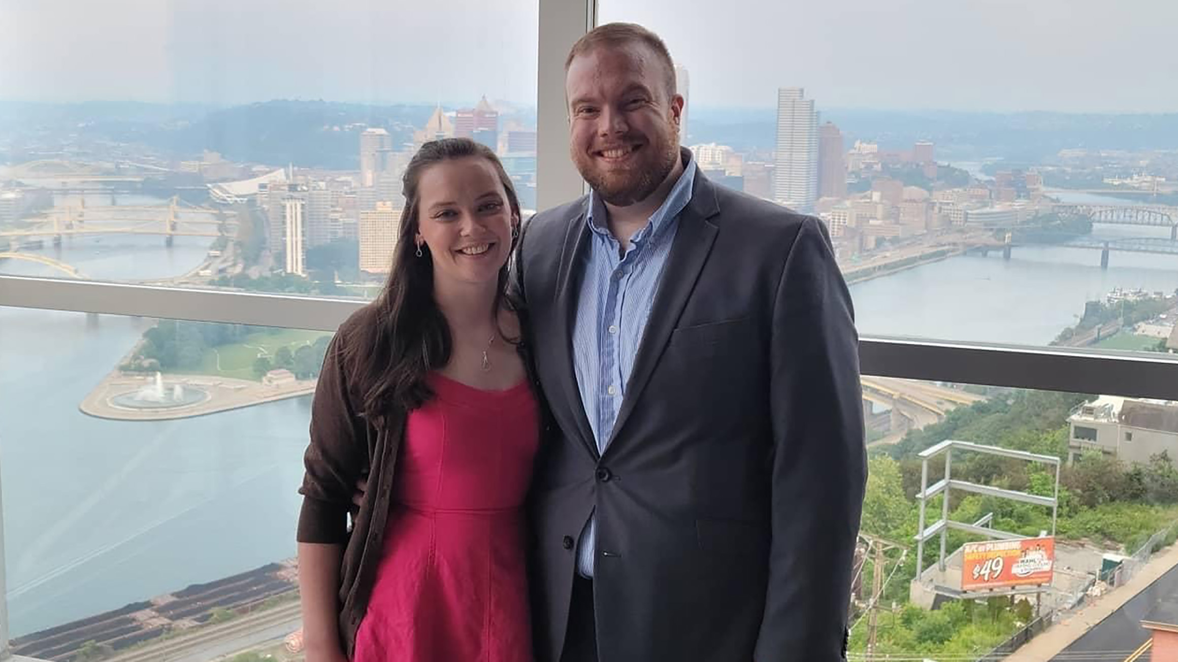 A man and woman pose in front of a window overlooking downtown Pittsburgh.