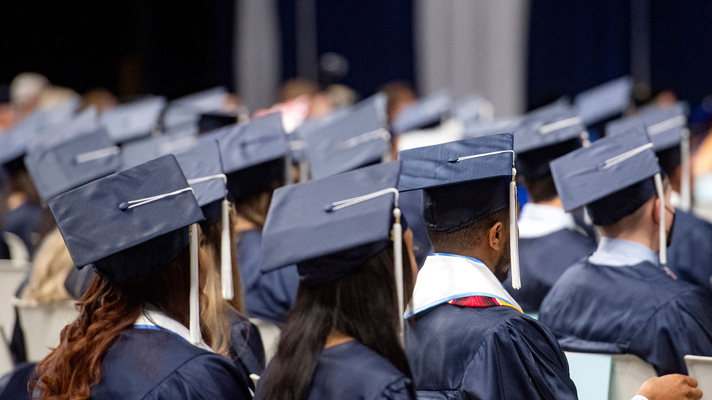 students wearing graduation caps and gowns sit in chairs at a commencement ceremony