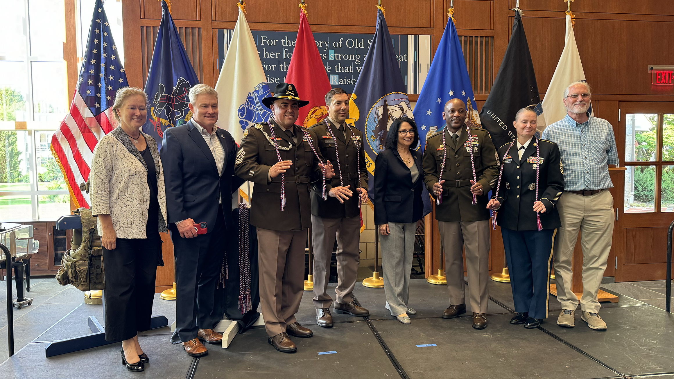 A group of people pose in front of the American flag and U.S. military flags.