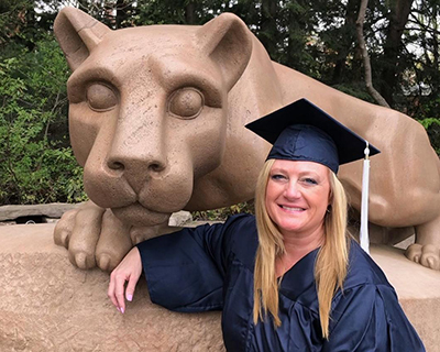 A woman wearing a blue graduation cap and gown poses in front of the Nittany Lion Shrine.