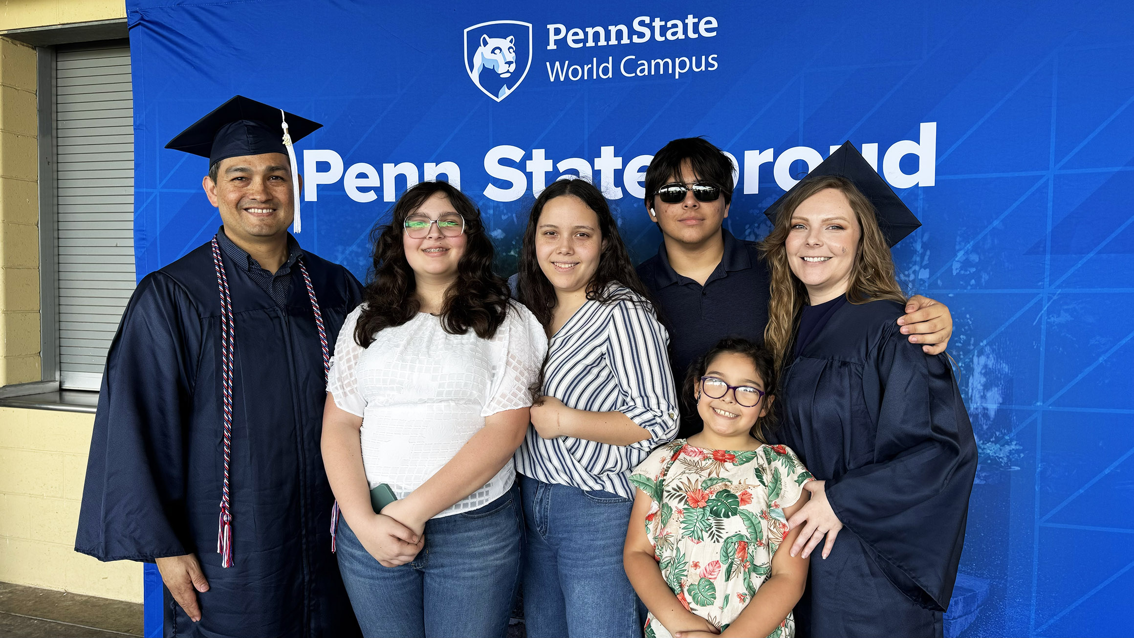 A man and a woman both wearing graduation caps and gowns pose with four children in front of a canvas that reads "Penn State Proud"