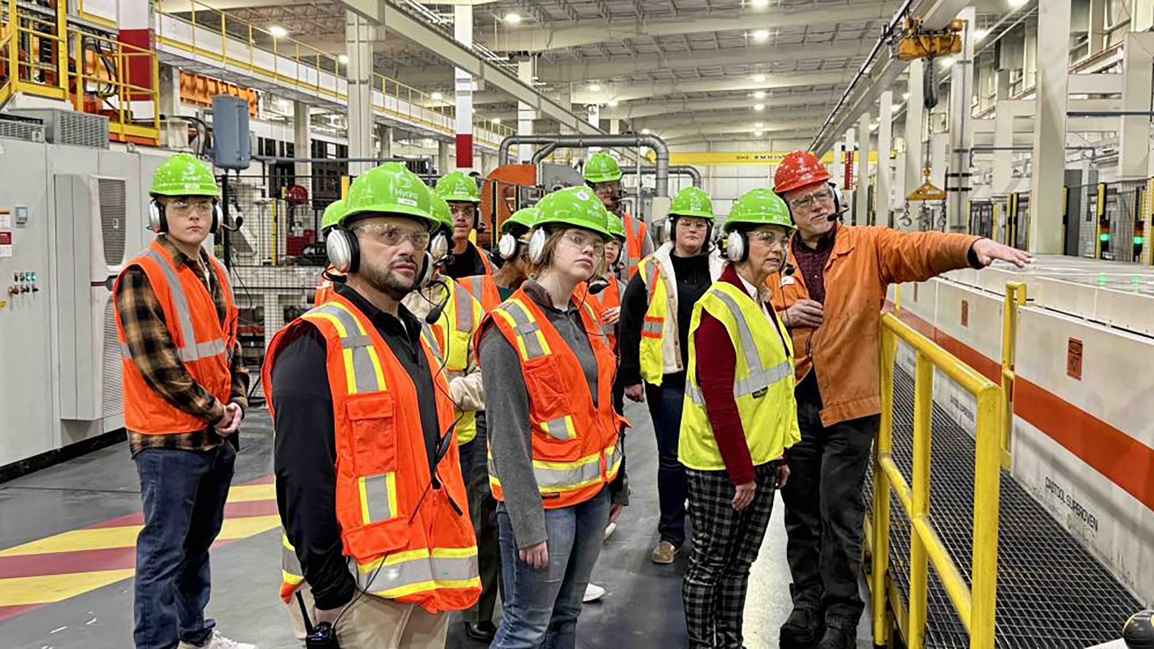 A group of people wearing reflective vests and hard hats stand in a factory.