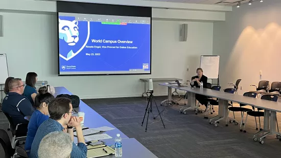 Renata Engel sits at a table and talks to students with a screen showing the title slide of her presentation. The title slide reads "World Campus Overview" and includes her title, which is vice provost for Online Education