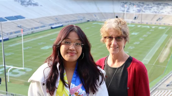 Two women pose in front of an empty football field and stadium.