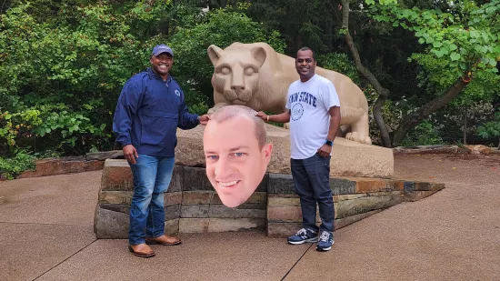 Two men pose with a large cutout of a man's head in front of the Nittany Lion Shrine.