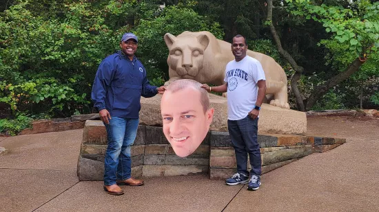 Two men pose with a large cutout of a man's face in front of the Nittany Lion Shrine.