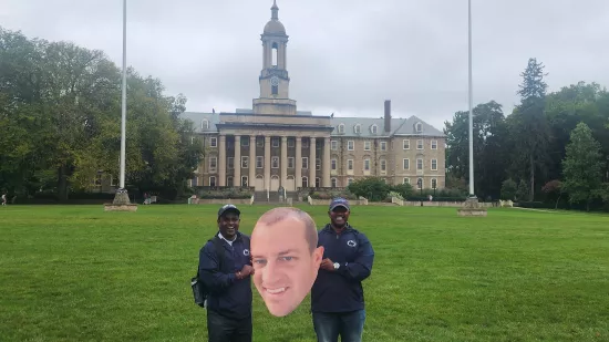 Two men pose with a large cutout of a man's face in front of Old Main.