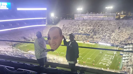 Two men pose with a large cutout of a man's face inside Beaver Stadium.