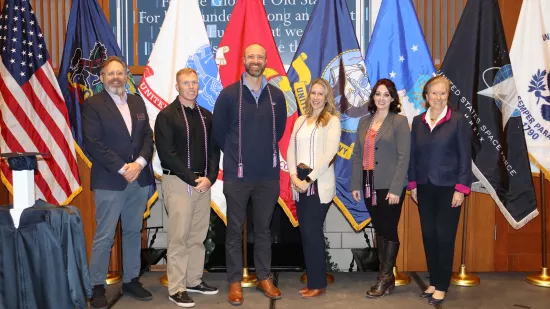 A group of people pose in front of the American flag and U.S. military flags.