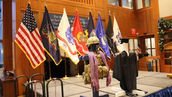 Military gear and a graduation cap and gown are displayed in front of the American flag and U.S. military flags.